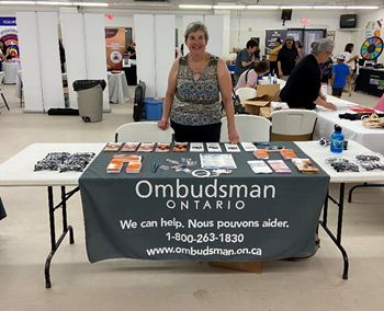 An Ombudsman Ontario staff member standing behind a table filled with Ombudsman giveaways and information materials, with a tablecloth that says “We can help.”